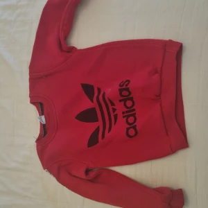 Barn kläder  - Säljer en snygg röd tröja från Adidas med det klassiska logotyptrycket i svart på framsidan förbarn 