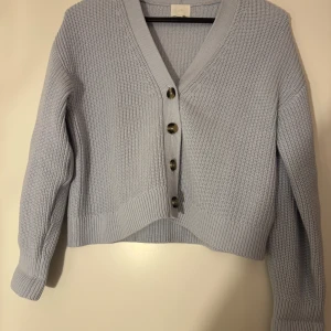 Ljusblå stickad kofta med knappar - Säljer en ljusblå stickad kofta från HM. Koftan har en v-ringning och stora knappar framtill. Den är långärmad med ribbade muddar och nederkant, vilket ger en mysig känsla. Perfekt för kyligare dagar!