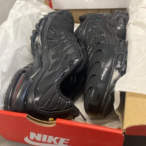 Svarta Nike TN - Snygga svarta Nike sneakers med en sportig design. Skorna har en luftig mesh-ovandel och en robust sula med synlig Air-dämpning för extra komfort. Perfekta för både vardag och träning.