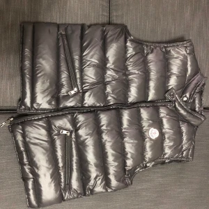 Svart dunväst från Moncler - Snygg svart dunväst från Moncler med dragkedja och två praktiska fickor framtill. Perfekt för kyligare dagar när du vill hålla stilen. Västen har en klassisk design med Moncler-logga på bröstet. Och priset går att diskutera 
