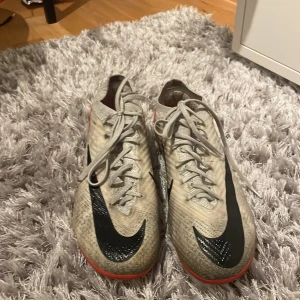 Nike fotbollsskor Mercurial - Säljer ett par Nike fotbollsskor i beige med svarta och röda detaljer. Skorna har snörning och en sportig design, perfekt för fotbollsplanen. De är i storlek herr 41 och har en stilren look med det klassiska Nike-märket på sidan. De har inte gått upp någonstans och påse kommer med.
