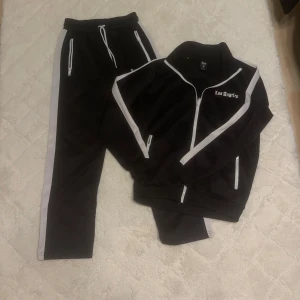 Svart träningsset med vita detaljer - Snyggt svart träningsset med vita detaljer och texten 'Los Angeles' på både jacka och byxor. Setet har dragkedjor på fickorna och en dragkedja framtill på jackan. Byxorna har en bekväm resårmidja med snörning.
