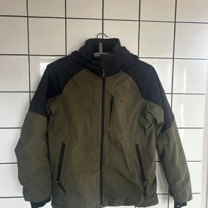 Grön och svart jacka från Everest - Säljer en grön och svart jacka från Everest med dragkedja framtill. Jackan har en huva och långa ärmar, perfekt för kyligare dagar. Den är tillverkad i ett slitstarkt material och har praktiska fickor med dragkedjor.