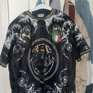 Svart fotbollströja med mönster - Snygg svart fotbollströja från Puma med ett detaljerat mönster och Italiens landslagsemblem på bröstet. Tröjan har korta ärmar och gulddetaljer som ger en exklusiv känsla. Perfekt för fotbollsfans som vill sticka ut.