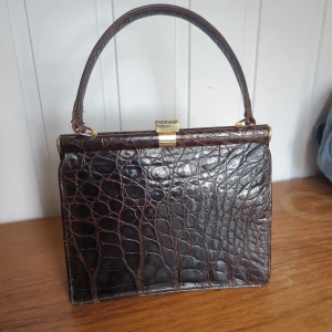 Retro ca 1950 skinn krokodilpräglad handväska - Retro, ca 1950 Elegant brun handväska med krokodilpräglat mönster och guldfärgad låsdetalj. Väskan har ett handtag och en dragkedja inuti för extra säkerhet. Perfekt för att ge en lyxig touch till din outfit.