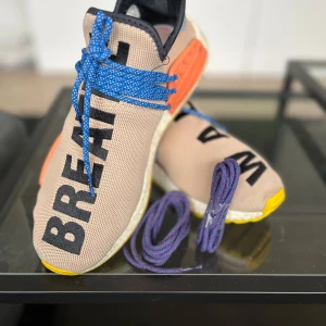 HUMAN RACE Adidas X Pharrell Wiliams  - Snygga sneakers från Adidas med beige ovandel och svart text. Skorna har blå snörning och en gul sula med mönster. Perfekta för en unik stil. Medföljer även extra snören i färgen lila som aldrig använts. Skick 3,5/5 original box finns ej 