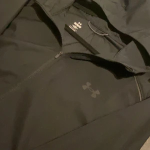 Svart vindjacka från Under Armour - Säljer en svart vindjacka från Under Armour med huva och dragkedja. Jackan har en stilren design med logotypdetaljer på bröstet och ärmen. Perfekt för blåsiga dagar och träning utomhus.