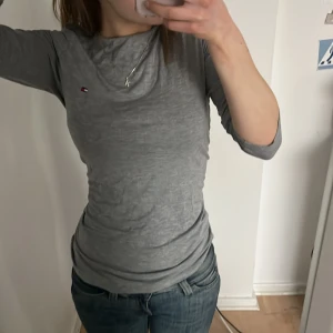 Grå långärmad tröja från Tommy Hilfiger - Säljer en stilren grå långärmad tröja från Tommy Hilfiger. Tröjan har en liten logotyp på bröstet och är perfekt för en avslappnad look. Passar bra till jeans eller kjol.