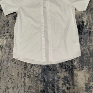 Vit kortärmad skjorta - Säljer en stilren vit kortärmad skjorta med knappar framtill. Perfekt för en avslappnad och fräsch look. Skjortan har en klassisk design som passar till många tillfällen.