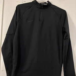 Svart långärmad tröja från Nike - Säljer en stilren svart långärmad tröja från Nike med dragkedja vid halsen. Perfekt för träning eller en avslappnad dag. Tröjan har en diskret Nike-logga på bröstet.