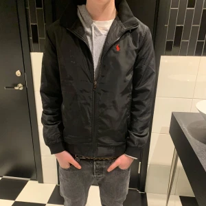 Svart jacka från Ralph Lauren  - Säljer en stilren svart jacka från Ralph Lauren med dragkedja och röd broderad logga på bröstet. Jackan har en klassisk design med ribbade muddar och krage. Perfekt för en casual look.
