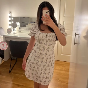 Blommig kortklänning - Charmig vit kortklänning med blommigt mönster och puffiga kortärmar. Klänningen har en söt rynkad detalj vid bysten och knytning i ryggen för en perfekt passform. Perfekt för sommarens alla tillfällen!