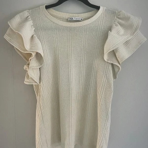 Beige ribbad topp från Zara - Säljer en stilren beige ribbad topp från Zara med volangdetaljer på ärmarna. 