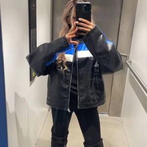 Svart och blå skinnjacka - Snygg svart skinnjacka med blå och vita detaljer. Jackan har en dragkedja framtill och en cool, oversized passform. Perfekt för en stilren look.