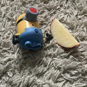 Minion-figur leksak - En liten Minion-figur med blå overall och en stor röd knapp på framsidan. Figuren har svarta handskar och skor, samt en grå detalj runt ögat. Perfekt för fans av de populära filmerna följer med äpple samskigt mums mums mums aah!!!