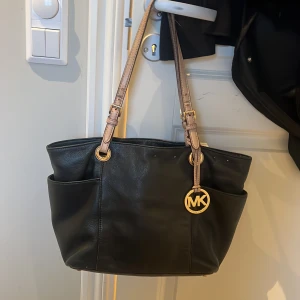 Äkta svart handväska från Michael Kors - Snygg svart handväska från Michael Kors med guldiga detaljer och bruna axelremmar. Väskan har flera praktiska fack och en stilren design. Perfekt för att bära med sig det viktigaste med stil. 