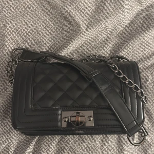 Svart quiltad väska med kedja - Snygg svart axelväska från NLY Accessories. Endast använd några få gånger. Liten defekt på framsidan men de är inget som märks (se sista bilden) säljer då den inte kommer till användning 💕 finns på vinted oxå!!