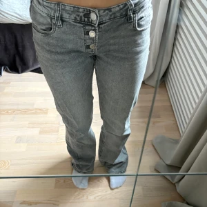 Grå jeans från Gina Tricot - Snygga grå jeans från Gina Tricot Young med låg midja och bootcut-stil. De har en knappgylf med flera knappar och klassiska femficksdetaljer. Perfekta för en trendig look!