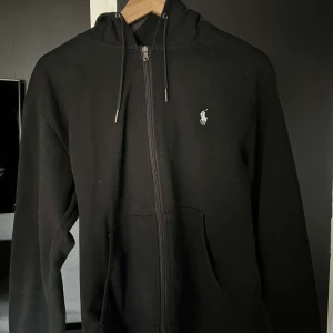 Svart hoodie från Ralph Lauren - Säljer en klassisk svart hoodie från Ralph Lauren med dragkedja och huva. Den har en liten broderad logga på bröstet och justerbara snören vid huvan. Perfekt för en avslappnad stil.