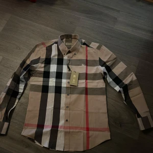 Rutig skjorta från Burberry - Snygg rutig skjorta från Burberry i beige, svart, vit och röd. Skjortan har långa ärmar och knappar framtill. Perfekt för en stilren look med klassiska rutor och en liten broderad detalj på bröstet. Storlek M men passar S