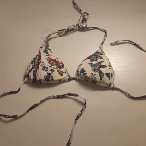 Blommig triangelbikinitopp med knytband - Jättefin bikinitopp med blommigt mönster i rött och blått på vit bakgrund. Bikinin har knytband både i nacken och ryggen för justerbar passform. 