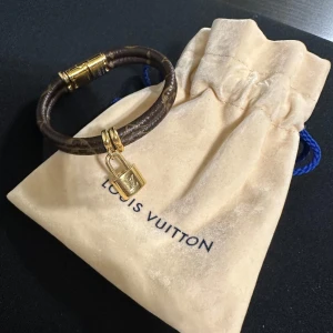 Brunt armband med lås från Louis Vuitton - Elegant brunt armband från Louis Vuitton med ett guldigt lås som hänge. Armbandet har ett klassiskt mönster och är tillverkat i ett slitstarkt material. Perfekt accessoar för att lyfta din stil. Säljs som på bild då jag kastat kartongen.