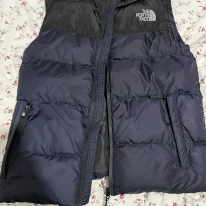Svart och blå dunväst från The North Face - Säljer en snygg svart och blå dunväst från The North Face. Västen har dragkedja och två praktiska fickor med dragkedjor. Perfekt för kyligare dagar och ger en stilren look.