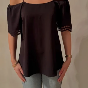 Svart offshoulder blus - Snygg svart offshoulder blus med spetsdetaljer på ärmarna. Perfekt för en stilren look. Blusen har en lös passform och korta ärmar.
