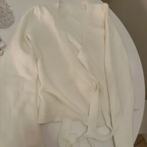 Vit omlottkofta - Säljer en stilren vit omlottkofta med långa ärmar och knytband i midjan. Perfekt för lager-på-lager och ger en elegant touch till din outfit. Passar bra till både vardag och mer uppklädda tillfällen.