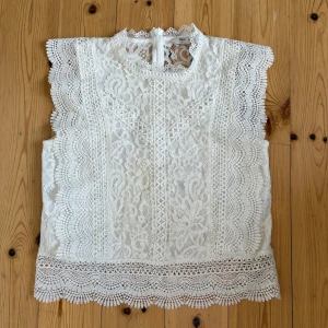 Vit spetsblus från ONLY - Säljer en elegant vit spetsblus från ONLY. Blusen är ärmlös med vackra spetsdetaljer och en hög hals. Perfekt för en stilren look. Det finns en liten svart fläck på framsidan!