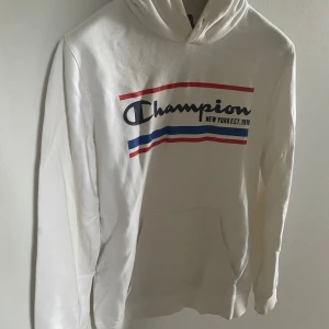 Vit hoodie från Champion - Vit hoodie från Champion med tryck i rött och blått på bröstet. Den har en klassisk känguruficka och Champion-logga på ärmen. 