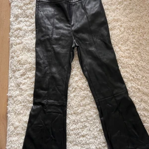Svarta fejkskinnbyxor från Nancy Cavally - Snygga svarta fejkskinnbyxor från Nancy Cavally. Byxorna har en bootcut-stil och har kort benlängd. Midjan har dragkedja i sidan. Perfekta för en trendig och high fashioned look. Storlek M