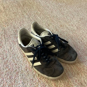 Adidas Gazelle i mörkblå mocka - Snygga Adidas Gazelle sneakers i mötkblå mocka med vita detaljer och klassiska tre ränder. Skorna har mörkblå snörning och en vit sula som ger en stilren look. Perfekta för en avslappnad stil.