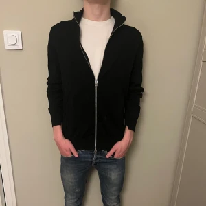 Svart helzip från j Lindberg  - Säljer denna riktigt feta helzip från j Lindberg i ett utmärkt skick som ni ser på bild 3 är det hål nere vid armarna vilket är design! Skriv vid fler frågor och funderingar. Plus ! Storlek m i tröjan men passar S ! 
