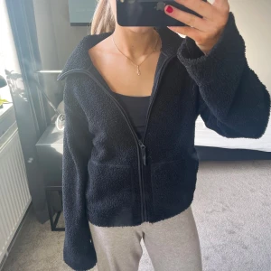 Svart teddyjacka med dragkedja - Mysig svart teddyjacka med dragkedja framtill och hög krage. Perfekt för kyliga dagar med sin fluffiga och varma design. Jackan har långa ärmar och en avslappnad passform.