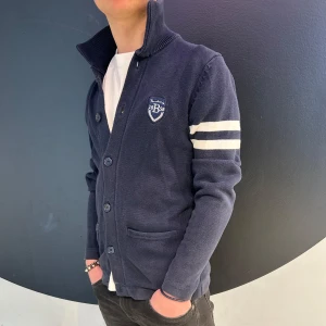 Bondelid, cardigan  - Snygg mörkblå kofta med knappar och ett stilrent emblem på bröstet. Två vita ränder på ärmen ger en sportig touch. Perfekt för en avslappnad stil.