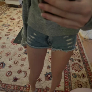 Blå jeansshorts med slitningar - Säljer ett par snygga blå jeansshorts med slitna detaljer och fransig kant. Perfekta för en avslappnad stil. De har en klassisk femficksdesign och knappgylf.