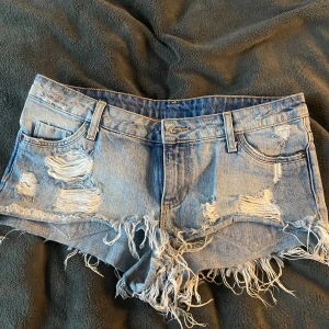 Lågmidjade jeansshorts  - Säljer ett par blå jeansshorts från H&m med slitna detaljer och fransar. Shortsen har en lågmidjad passform och klassisk femficksdesign. Perfekta för en sommaren. De är även väldigt små i storleken så skulle säga att de lätt passar en S/36