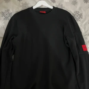 Svart långärmad tröja från Hugo Boss - Snygg svart långärmad tröja från Hugo Boss med en röd detalj på ärmen. Perfekt för en stilren look. Tröjan har en klassisk passform och är tillverkad i ett mjukt material.Pris kan diskuteras!!