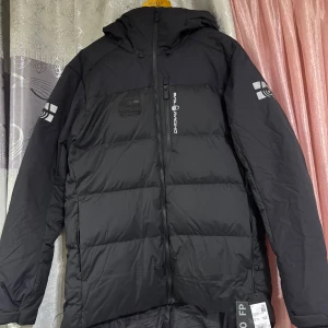 Svart dunjacka från Sail Racing - Säljer en svart dunjacka från Sail Racing i storlek XL. Jackan har en stilren design med dragkedja och huva. Perfekt för kyliga dagar med sin varma dunfyllning. Märkesdetaljer på ärmarna och bröstet ger en sportig look.