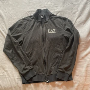 Svart jacka från Emporio Armani - Säljer en stilren svart jacka från Emporio Armani med dragkedja framtill. Jackan har en normal passform och är långärmad. Perfekt för en casual look med sin enkla design och diskreta logga på bröstet. Äkta 