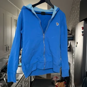 Blå hoodie från Paul Smith - Snygg blå hoodie från Paul Smith med dragkedja och en färgglad broderad zebra på bröstet. Den har en ljusblå insida i huvan och långa ärmar. Perfekt för en avslappnad stil.