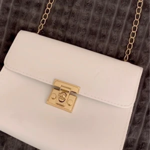 Vit axelväska med guldkedja - Snygg vit axelväska med en elegant guldkedja och ett stilrent lås framtill. Perfekt för att ge din outfit en touch av lyx. Väskan har en enkel och chic design som passar till många tillfällen.
