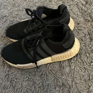 Svarta Adidas NMD sneakers - Säljer ett par svarta Adidas NMD sneakers med vit sula och klassiska tre ränder på sidan. Skorna har en stickad ovandel och snörning för en bekväm passform. 