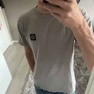 Grå t-shirt från Stone Island Junior - Säljer en stilren grå t-shirt från Stone Island Junior med deras ikoniska logga på bröstet. Perfekt för en avslappnad look. T-shirten har en klassisk passform och är kortärmad. Mycket bra skick. Passar mig som är 182 och väger 60kg. Ny pris runt 1300. Mitt pris 550