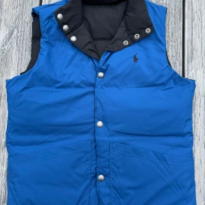 Blå och svart dunväst från Ralph Lauren barn - Säljer en snygg och vändbar dunväst från Ralph Lauren i blått och svart. Västen har tryckknappar och en stilren design med märkets logga på bröstet. Perfekt för kyligare dagar!