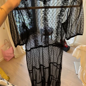 Svart transparent klänning med prickar - Snygg svart långklänning med prickigt mönster och korta ärmar. Den har volangdetaljer och en svart underklänning med smala axelband.