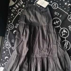 Svart långkjol med volanger - Snygg svart långkjol med volanger som ger en härlig rörelse. Kjolen har en elastisk midja för bekväm passform. Perfekt för att skapa en stilren look. Helt ny med prislappen kvar. 