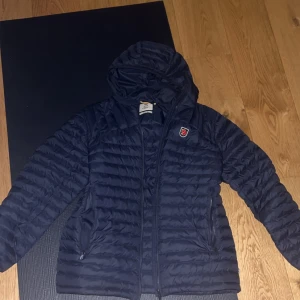 Blå Fjällräven Expedition Jacket - Säljer en blå Fjällräven Expedition Jacket med huva och dragkedja. Jackan är quiltad och har en klassisk design med Fjällrävens logga på bröstet. Priset är inte fast än.