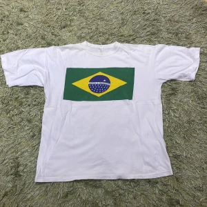 Vit t-shirt med brasiliansk flagga - Vit t-shirt med tryck av den brasilianska flaggan på framsidan. Perfekt för att visa ditt stöd för Brasilien! T-shirten har en klassisk passform och korta ärmar.